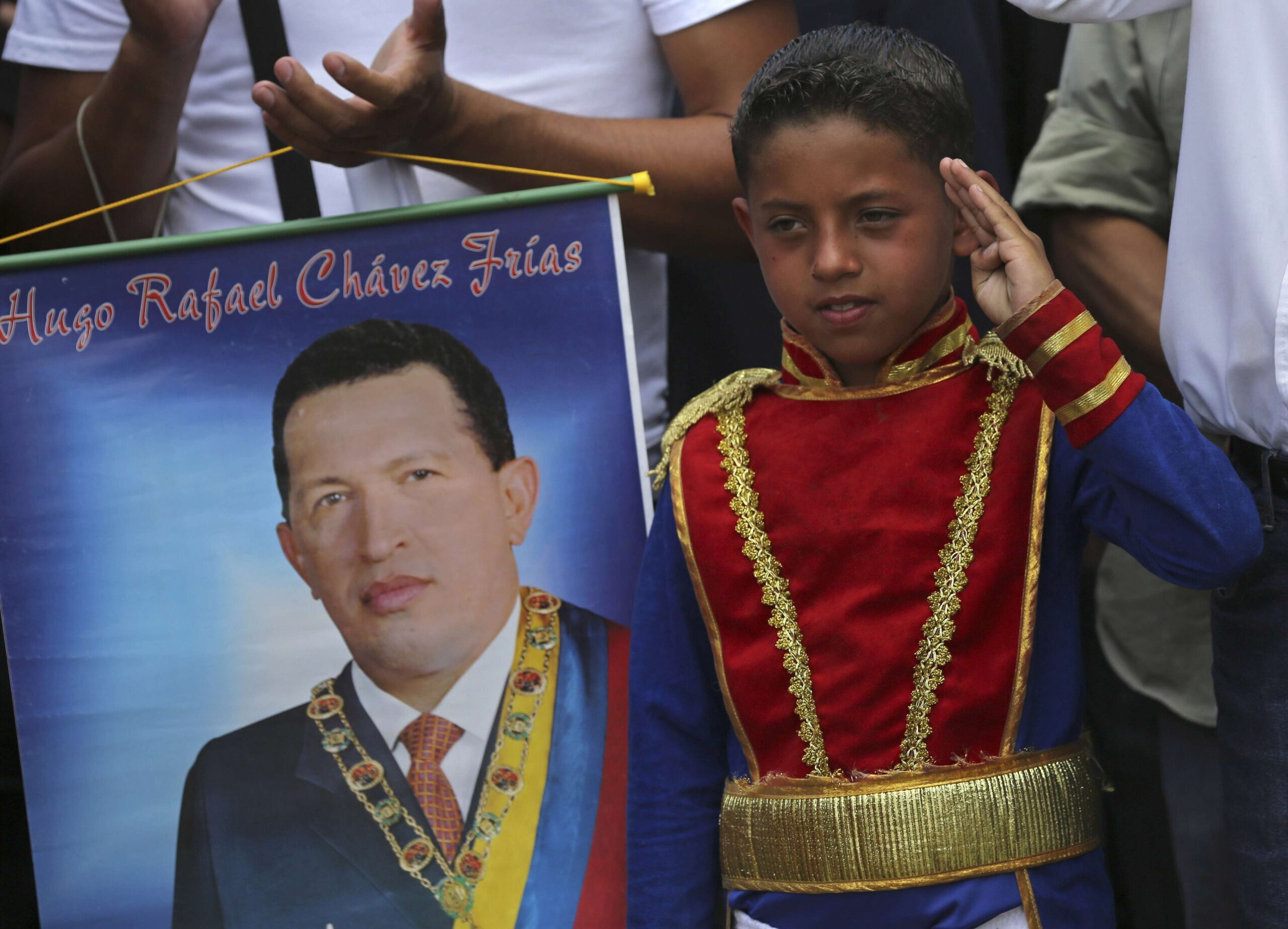 Un niño vestido con un uniforme histórico asociado con el héroe venezolano Simón Bolívar saluda mientras está de pie junto a un cartel del fallecido presidente Hugo Chávez durante un desfile militar en conmemoración del primer aniversario de la muerte de Chávez, en Caracas, Venezuela, miércoles 5 de marzo de 2014. © AP Foto/Fernando Llano