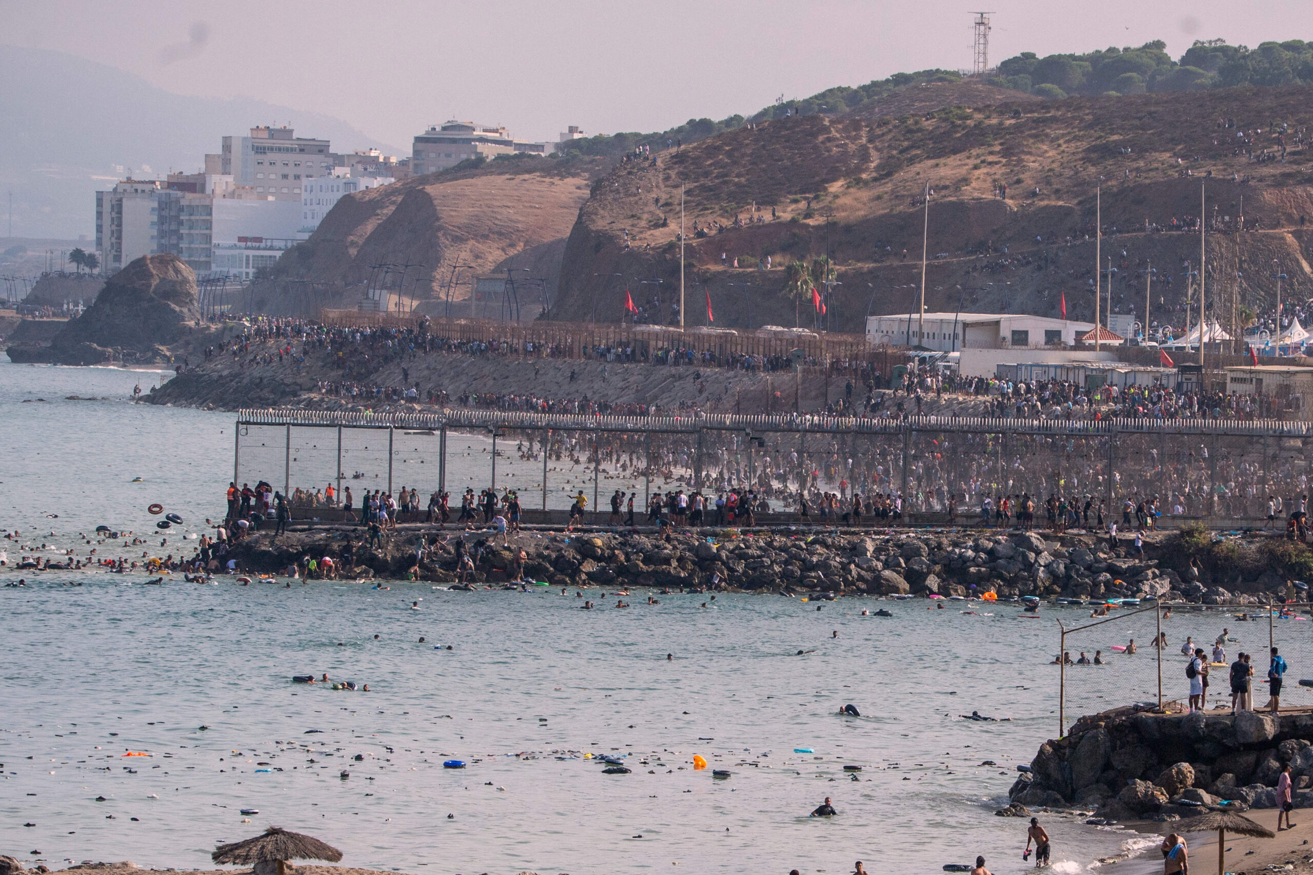 A lo largo de la valla de Tarajal, miles de personas bajan de las colinas hacia la costa por los diques de rocas, bajo la mirada de los agentes españoles apostados en el muelle, el jueves 30 de julio de 2026. © AP Photo/Antonio Sempere/SIPA