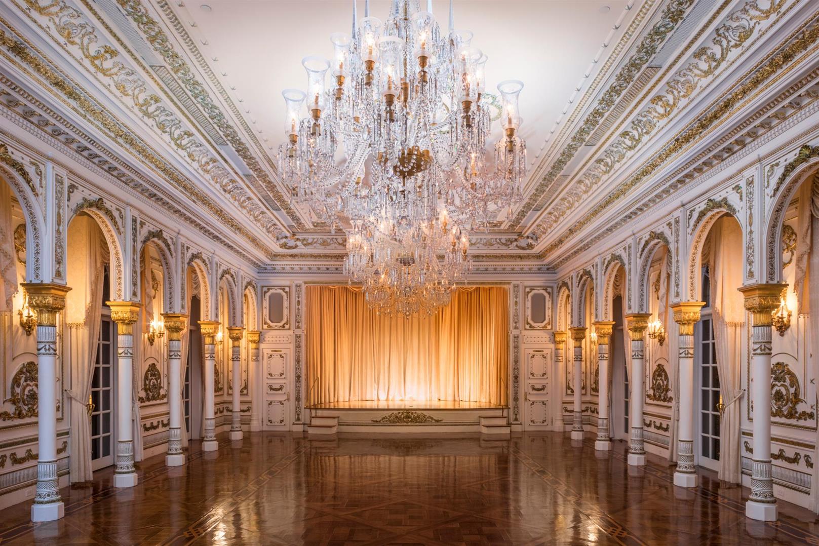 Uno de los salones de recepción de Mar-a-Lago: lámparas de cristal en cascada, columnas doradas y un escenario con telón dorado.