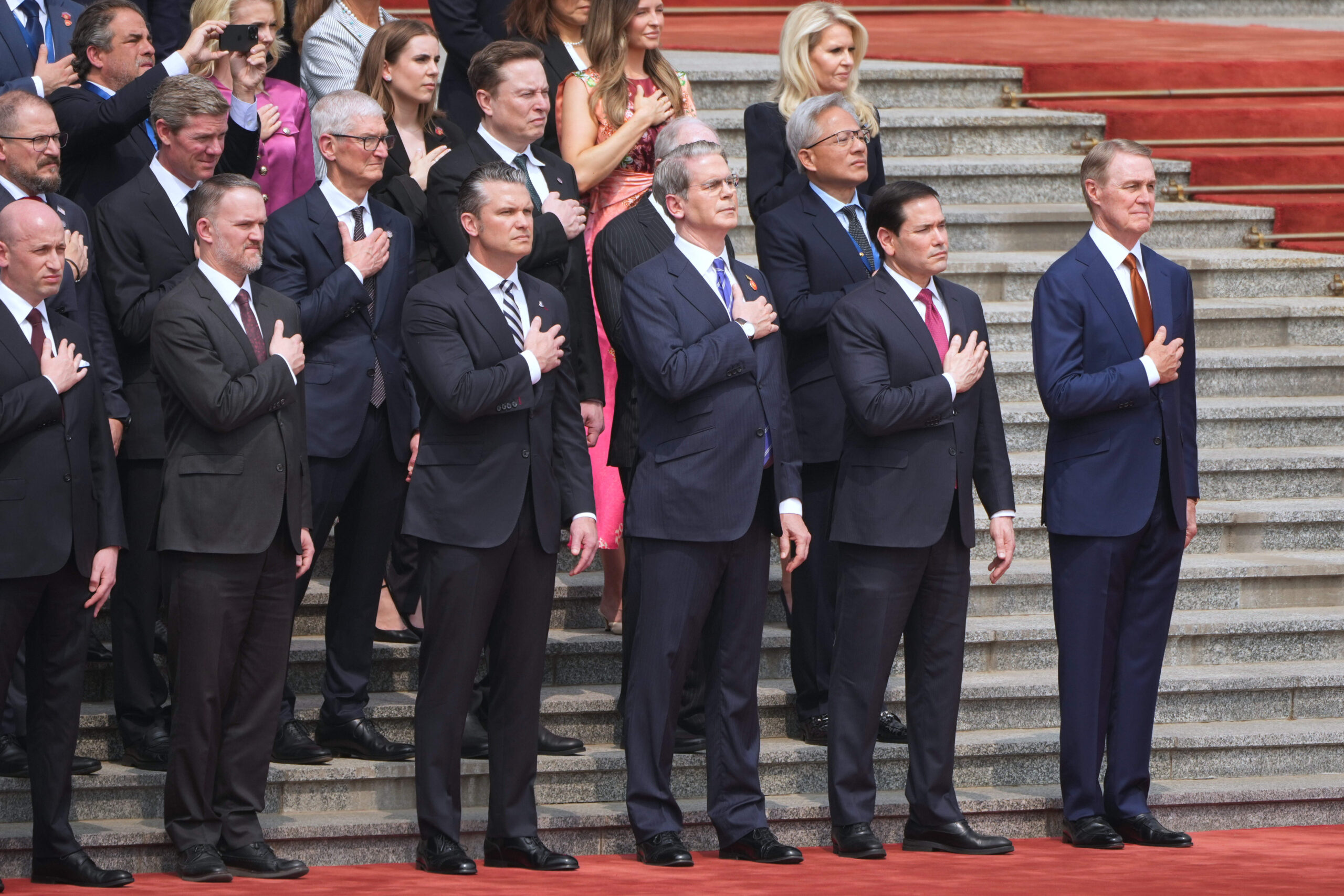 Les membres de la délégation américaine, parmi lesquels Stephen Miller, chef de cabinet adjoint de la Maison Blanche Jamieson Greer, représentant américain au Commerce, Pete Hegseth, secrétaire à la Guerre, Scott Bessent, secrétaire au Trésor, et Marco Rubio, secrétaire d’État, l'ambassadeur des États-Unis en Chine David Perdue, le PDG d'Apple Tim Cook, Elon Musk et le PDG de Nvidia Jensen Huang, écoutent l'hymne national alors que le président Donald Trump participe à la cérémonie de bienvenue. © AP Photo/Mark Schiefelbein