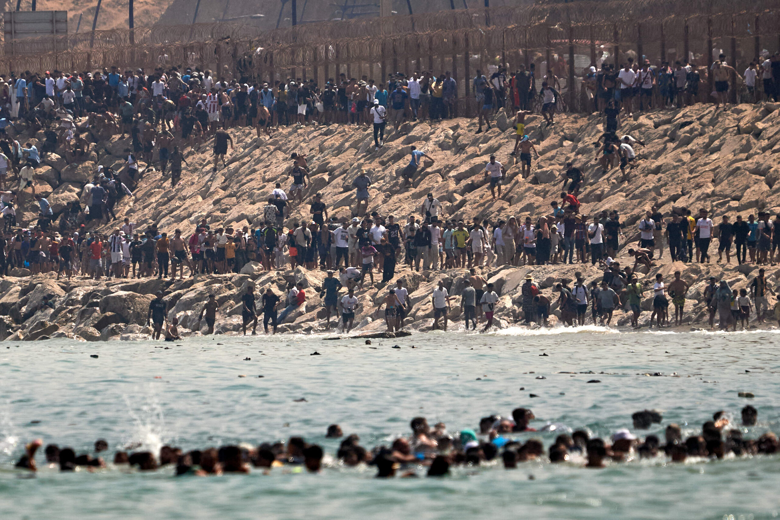 Le long de la clôture du Tarajal, des milliers de personnes descendent des collines vers le rivage par les enrochements, sous le regard des agents espagnols postés sur la jetée, jeudi 30 juillet 2026. © AP Photo/Antonio Sempere/SIPA