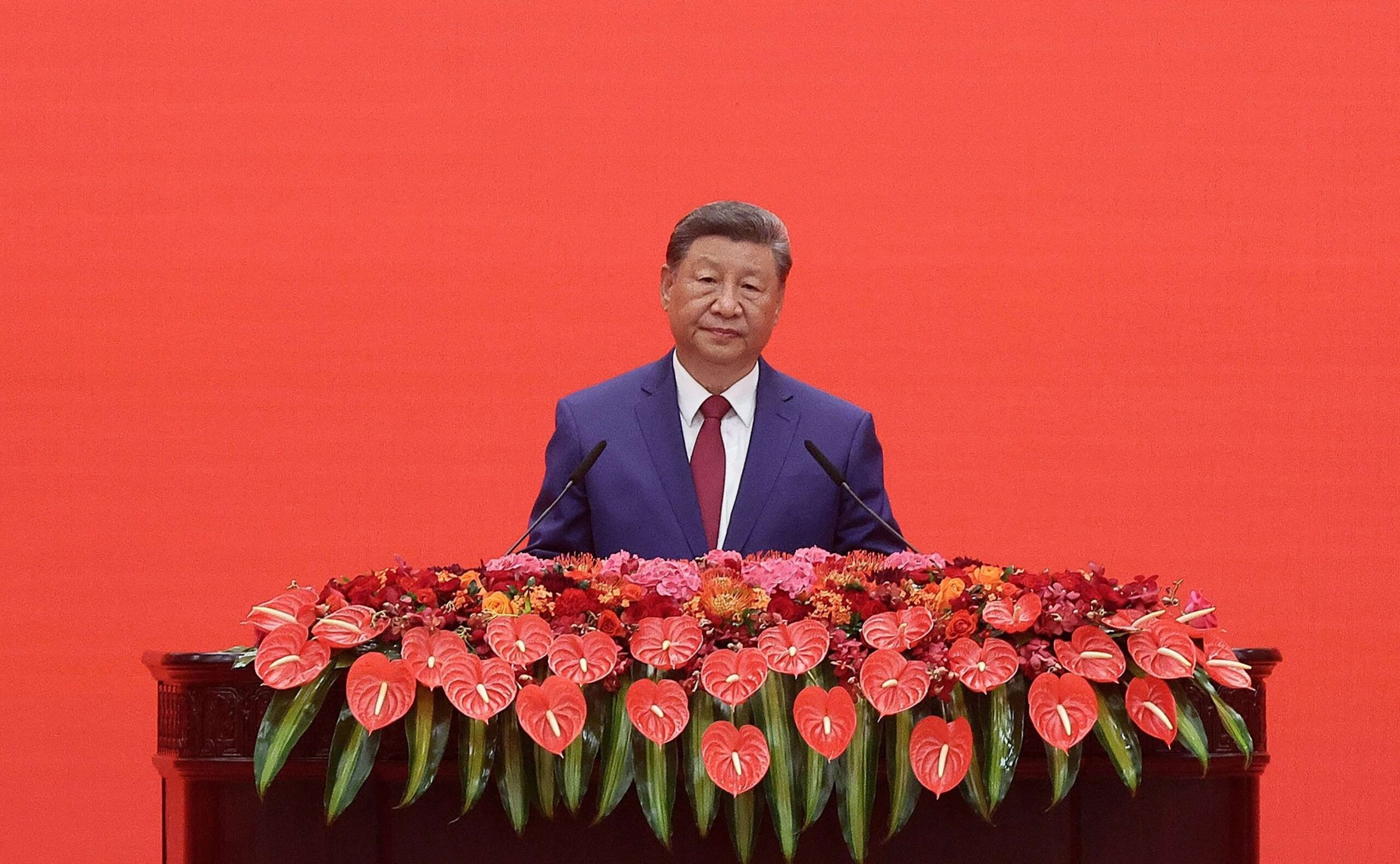 Face aux convives, Xi au pupitre sur fond rouge uni, derrière une cascade d'anthuriums en forme de cœur : la scénographie remplace les missiles du matin par des fleurs, même rigueur, autre registre. Le message aussi change d'habit : la puissance de Tian'anmen se présente au banquet en garant de la paix.  Photo : Vladimir Smirnov / TASS