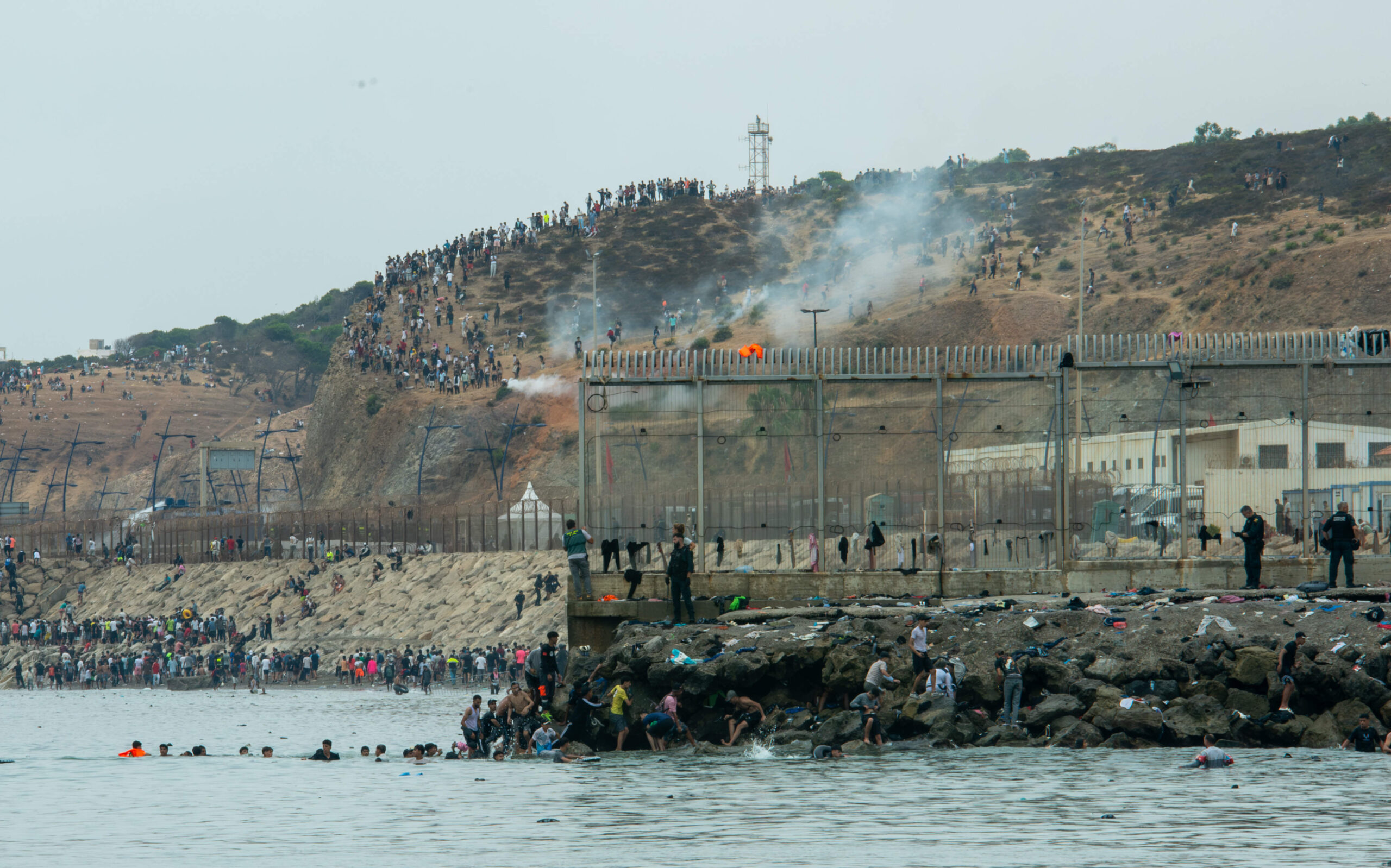 Des milliers de personnes sur la colline, des nageurs sont dans l'eau, des vêtements sont abandonnés sur les barbelés, le 31 juillet 2026, dans les fumées de gaz lacrymogène. Photo : Juan Carlos Lucas